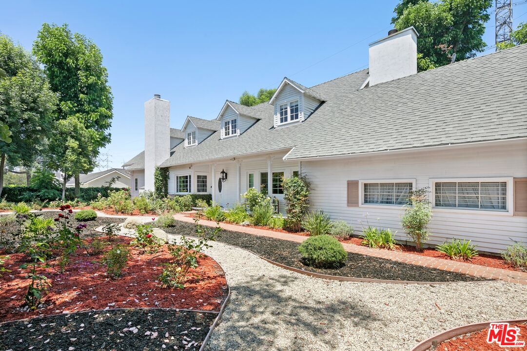 White house with multiple dormers and chimneys, set behind landscaped gardens and curving paths.