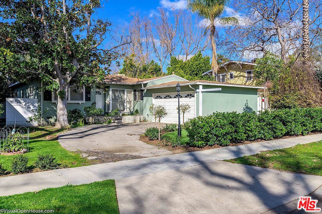 Single-story green house with an attached garage, broad driveway, and trimmed front hedges.